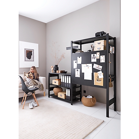 Une femme lit sur un canapé. Étagère noire avec notes et décor. Tapis clair, chaise.