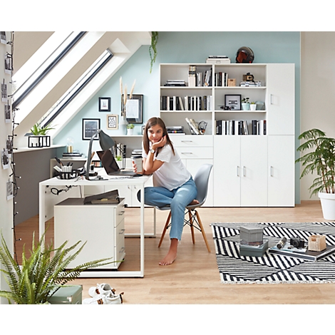 Jeune femme assise pieds nus à un bureau avec un ordinateur portable. Dans l'appartement mansardé se trouvent une armoire, une bibliothèque et des plantes en pot.