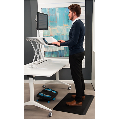Man stands at a height-adjustable desk with laptop, monitor and footrest on a mat.