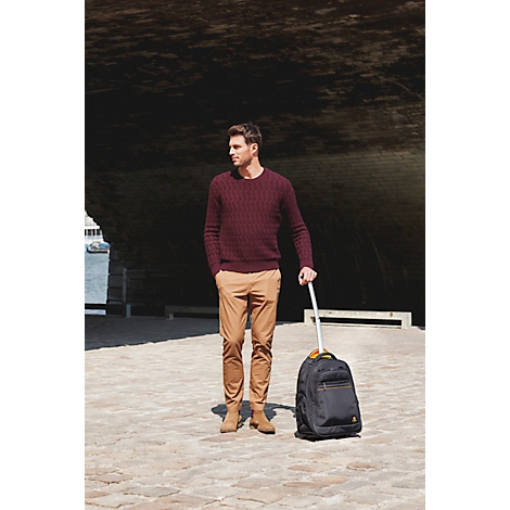 Ein Mann in weinrotem Pullover und beigefarbener Hose steht mit Rollkoffer. Er blickt zur Seite.
