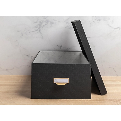 Schwarze, geöffnete Aufbewahrungsbox mit Deckel. Mit weißem Etikett und goldenem Griff auf Holz. Marmorhintergrund.