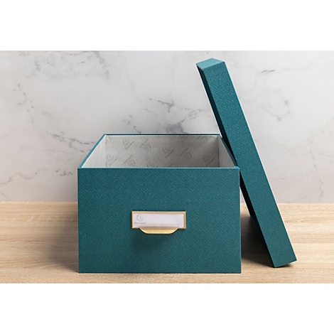 Une boîte teal ouverte avec un couvercle, posée sur une table en bois. Une étiquette avec une poignée est attachée à la boîte.