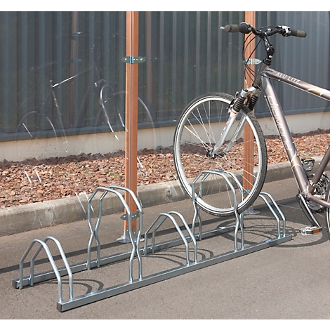 Fahrrad steht in einem Fahrradständer. Hinter dem Ständer befindet sich eine durchsichtige Wand und braune Stützen.