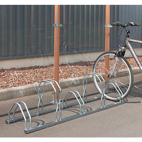 Fahrradständer aus Metall mit Fahrrad davor. Im Hintergrund eine transparente Wand und eine graue Fassade.