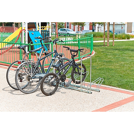 Drei Fahrräder stehen auf einem Gehweg in einem Fahrradständer. Im Hintergrund befindet sich ein Spielplatz.
