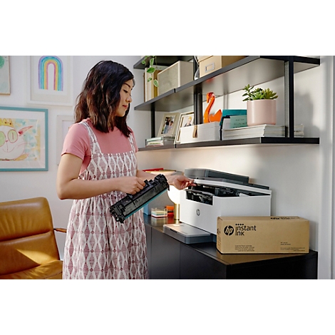 Eine Frau wechselt in ihrem Büro eine Druckerpatrone. Auf dem Tisch steht ein Drucker. Auf der Box steht: HP Instant Ink.