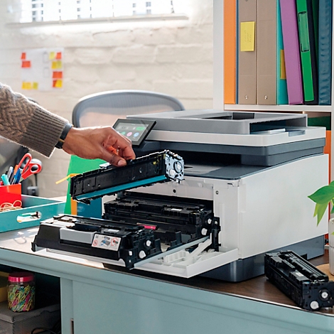 Person tauscht Tonerpatrone in einem weißen Drucker. Die Schublade ist geöffnet. Bücherregal im Hintergrund.