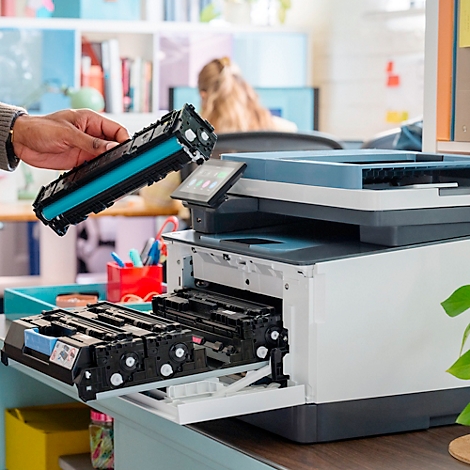 Eine Hand hält eine blaue Tonerkartusche über einem Drucker. Eine Person ist im Hintergrund unscharf zu sehen.