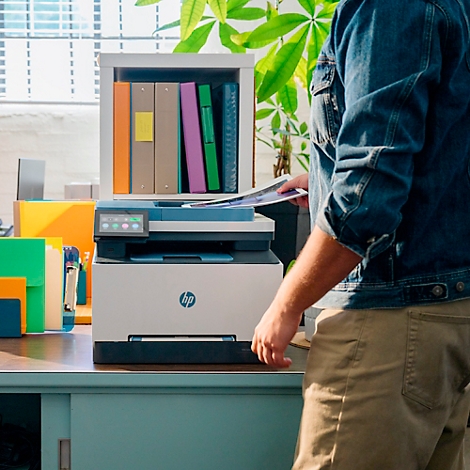 Eine Person legt Papier in einen HP-Drucker. Der Drucker steht auf einem Schreibtisch, im Hintergrund sind Ordner und Pflanzen zu sehen.