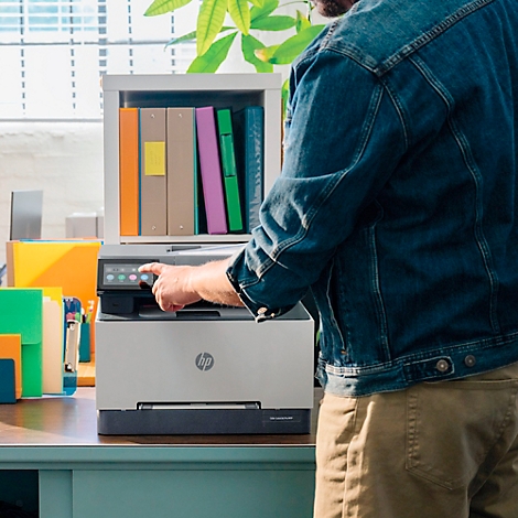 Eine Person legt Papier in einen weißen Drucker der Marke HP. Im Hintergrund ein Schreibtisch mit Ordnern und Büchern.