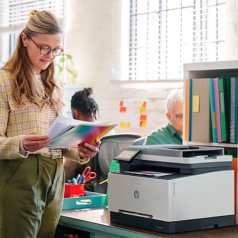 Eine Frau betrachtet Dokumente neben einem Drucker. Im Hintergrund: Büro-Mitarbeiter. HP-Drucker stehen auf einem Schreibtisch.