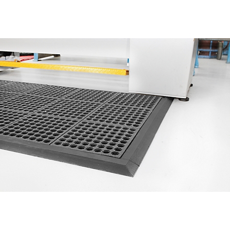 Black workstation mat with rounded edges and hole pattern. It lies in front of a machine on a white floor.