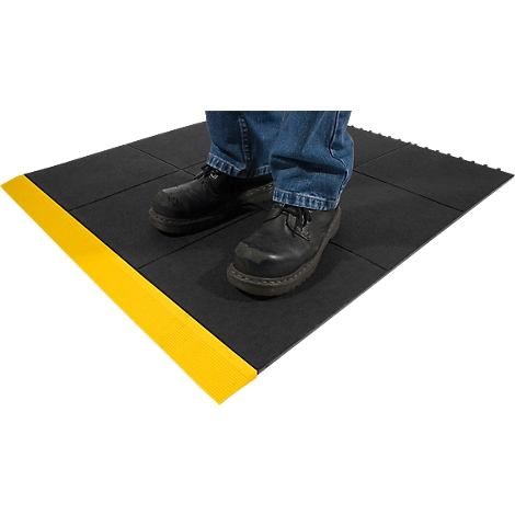 A person stands on a black anti-slip mat with a yellow edge. Feet are in black shoes and jeans.