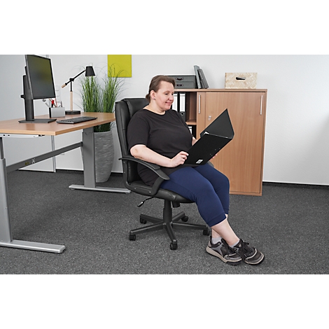 Femme assise dans un fauteuil de bureau, lisant un dossier. Elle porte un haut noir et un pantalon capri bleu. Bureau et armoire en arrière-plan.