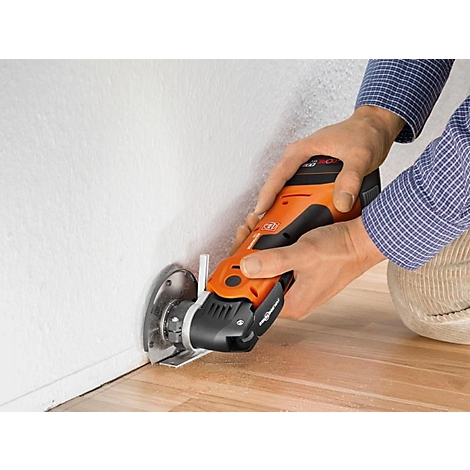 Une personne coupe dans un mur avec un multi-outil orange et noir. Du bois sur le sol.