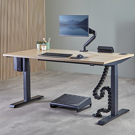 Ein höhenverstellbarer Schreibtisch mit Monitor, Tastatur, Maus und Fußstütze. Holzoptik, graue Beine, grüner Hintergrund.