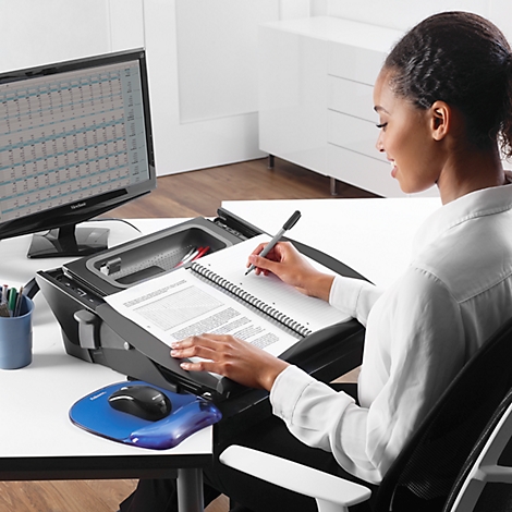 A woman writing in a notebook. She sits at a desk with a monitor and a stand that contains documents and a mouse.