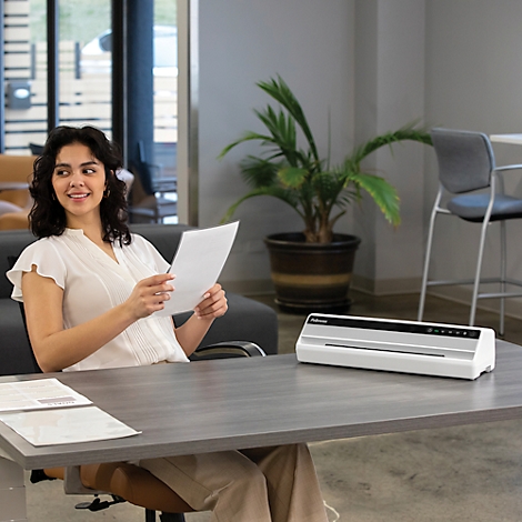 A smiling woman holds paper, a laminator sits on a desk.