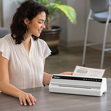 A smiling woman laminates a sheet with the word "GOALS". A white laminator is on the table.