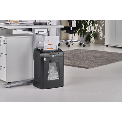 A black paper shredder stands next to a desk. Paper is being shredded at the top, and the container below is filled with shredded paper.