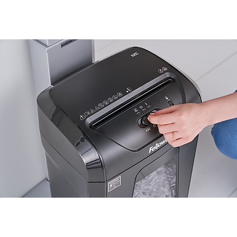 A person operates a black paper shredder. The hand is turning a knob. Shredded paper is already in the collection bin.