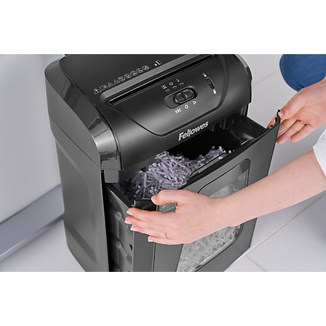 Person opening the waste bin of a black paper shredder. The bin is filled with shredded paper. "Fellowes" is visible on the device.