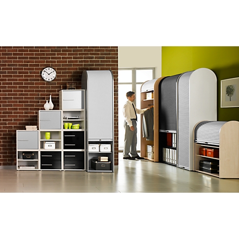 Man in office at cabinet wall, shelves to the left. Brick wall, clock, green wall.