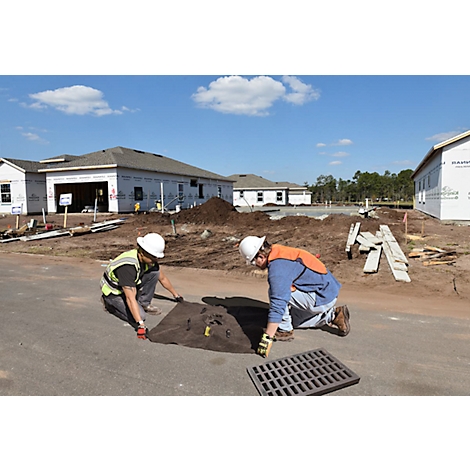 Deux ouvriers du bâtiment agenouillés sur le bord d'une route travaillant sur un drain. Des maisons en construction sont visibles en arrière-plan.