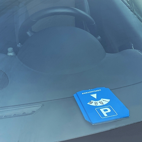 Blauer Parkausweis auf der Windschutzscheibe eines Autos. Der Ausweis zeigt die Ankunftszeit an. Im Hintergrund das Lenkrad.