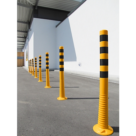 Line of yellow bollards with black stripes. On asphalt in front of a white wall, a facade with a roof on the left.