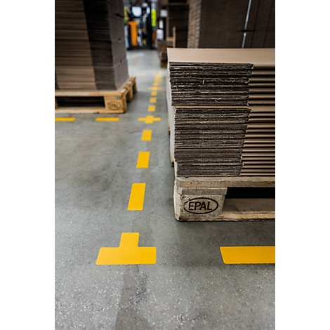 Yellow markings on a concrete floor, stacks of cardboard on pallets. Forklift and other goods in the background.