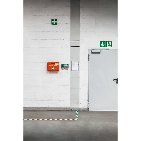 Interior shot with a white wall, featuring a first aid kit, a green cross, and an emergency exit sign. A green and white tape is on the floor.