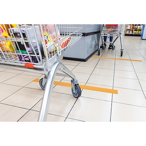 Shopping cart filled with groceries stands in front of a checkout counter. Yellow line on the floor marks distance.