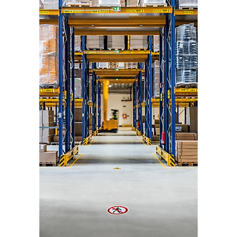 Warehouse with high shelves. A forklift truck drives in the distance. A sign with a prohibited footpath is on the floor.