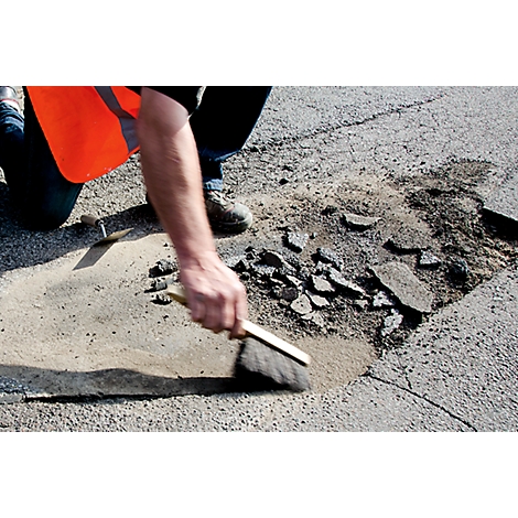 A person sweeps a road that has a pothole. The worker wears an orange safety vest.