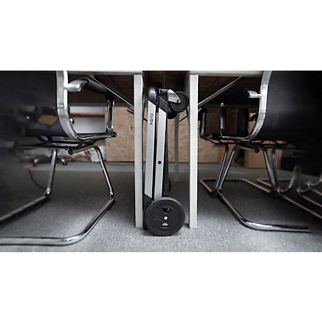 A folded walker stands under a table, between chairs. It is grey and black, with two wheels.