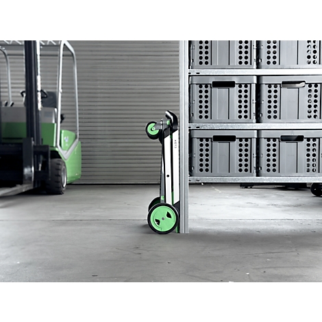 Foldable shopping trolley with green wheels stands next to a shelf with grey boxes and a forklift.