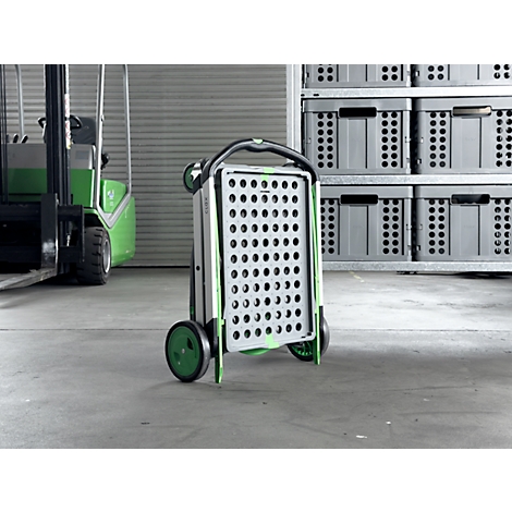 A folded gray shopping trolley with green wheels stands in a warehouse, a forklift and gray boxes in the background.