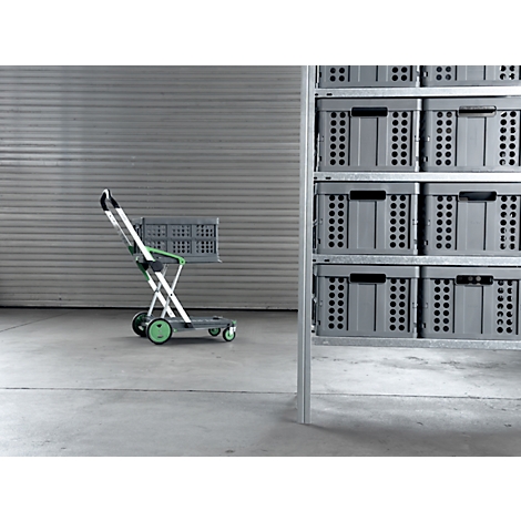 Grey shopping trolley with basket, in front of shelf with boxes. Green accents on wheels and handle. On concrete floor in front of corrugated wall.