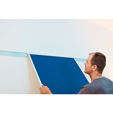 A man attaches a blue wall panel to a rail on the wall. He is looking at the panel.