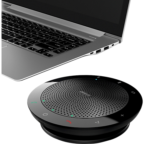 Ein schwarzes Konferenzsystem und ein silberfarbener Laptop liegen nebeneinander. Das Konferenzsystem ist rund, mit Bedienelementen und einem perforierten Lautsprecher.