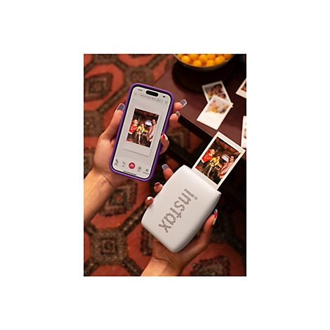 Person hält ein Smartphone mit Foto und einen weißen Instax-Drucker, aus dem ein Foto kommt. Bunte Fingernägel.