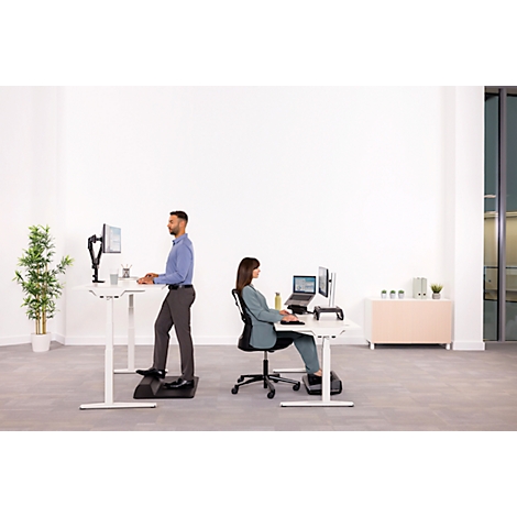 Ein Mann steht an einem höhenverstellbaren Schreibtisch, eine Frau sitzt. Beide arbeiten an Computern in einem hellen Büro.