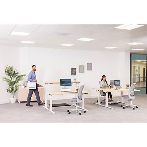 Büro mit zwei höhenverstellbaren Schreibtischen und Mitarbeitern. Ein Mann geht, eine Frau sitzt. Helle, moderne Atmosphäre.