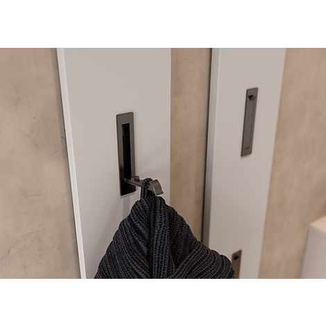 Weißes Wandpaneel mit Haken und einem grauen Pullover, an einer beigen Wand. Drei dunkle, längliche Vertiefungen.