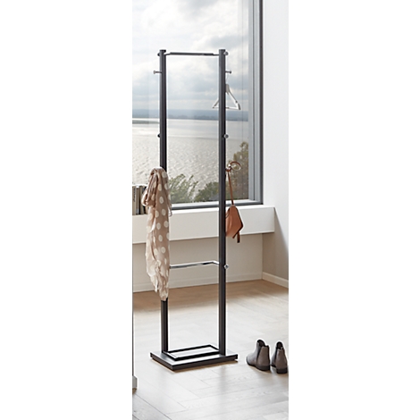 Schwarze Garderobe mit Kleiderbügeln, Schal und Tasche. Graue Schuhe stehen davor. Im Hintergrund ist eine Fensterfront mit Blick auf einen See.