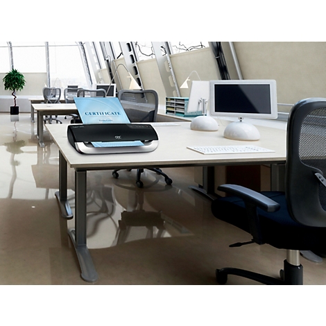 Ein schwarzer Drucker auf einem Schreibtisch, ein Zertifikat wird ausgedruckt. Büro-Szene mit Computer und Pflanzen.