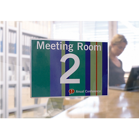 Ein Schild mit der Aufschrift "Meeting Room 2". Im Hintergrund eine Frau an einem Schreibtisch.