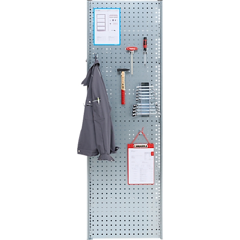 Gereedschapsmuur met gereedschap, klembord en jas. Metalen constructie, lichtgrijs. Diverse gereedschappen aan de muur bevestigd.