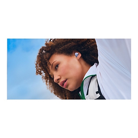 Nahaufnahme einer Frau mit Locken und Ohrhörern. Sie blickt nach unten, vor blauem Himmel und mit weißer Jacke.